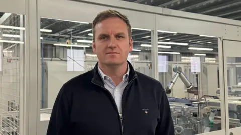 Cormac Diamond of Bloc Blinds has short brown hair and is staring into the camera. He is wearing a patterned shirt underneath a dark jacket. In the background are machines used to make blinds.