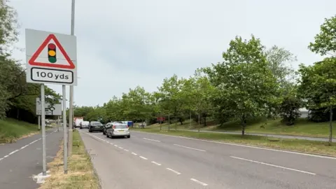 Jamie Niblock/BBC A general view of Uppermayne, which is a dual carriageway road with trees separating traffic in either direction. Cars are queuing for a roundabout. 