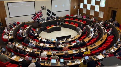 The meeting room at County Hall is full of councillors. It has seats arranged in a circle with desks in front of them angled around a singular desk. There is a UK flag and a St Piran flag crossed at the front of the hall and two large monitors behind them.