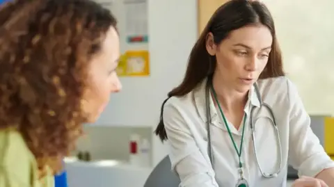 A photograph of a female GP with a stethoscope around her neck talking to her patient who is also a woman 