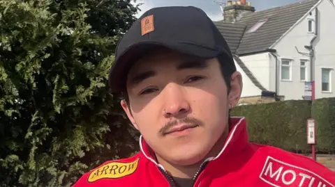A young man in his 20s sat on a park bench. He is wearing a red-and-white retro Formula 1 tracksuit and a baseball cap. He also has a small pencil moustache.