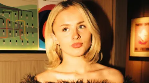 Gracie Hall Mel Raeburn, a woman with long blonde hair and wearing a black furry top, stands against a wall while looking to her right.