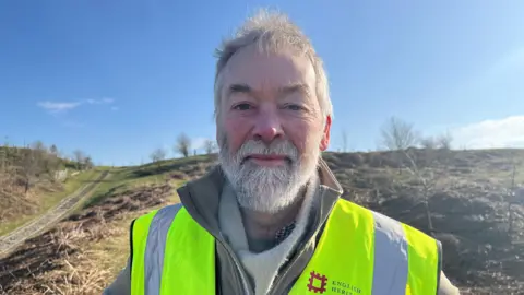 Ian pictured on one of the slopes of the hillfort. He's wearing a khaki waterproof coat, with a yellow hi-vis vest over the top. On his left lapel the vest has the English Heritage name and logo (a red square shaped like a portcullis). Behind him are grassy slopes covered in bracken and small trees. The sky is bright blue with bright sunshine. 