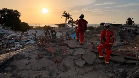 Mark Leeson Two people are in orange firefighter suits with backpacks and helmets on. They are walking over a large pile of rubble that looks to be concrete and brick. In the distance is an orange and yellow sunset above the ocean.