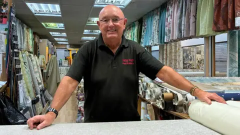 A man wearing glasses and a black polo shirt with Billy Hill Curtains written on is smiling while standing at his stall. He is surrounded by curtain samples.