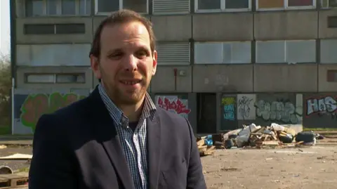 A man standing in front of a block of flats with graffiti on its walls. There is a pile of rubbish in front of the building. The man has short brown hair and facial hair and is wearing a black suit jacket with a blue and white chequered shirt open at the collar.