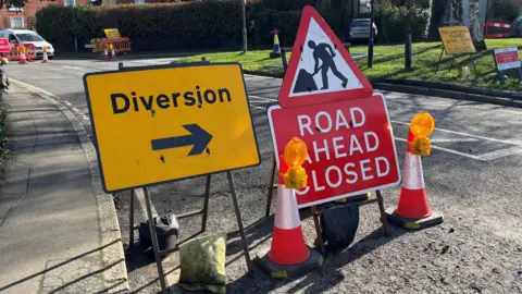 Jodie Halford/BBC Two road signs, one is yellow that reads 'Yellow' with a sign pointing to the left and the other one says Road ahead closed.
There are two traffic cones also placed nearby. 