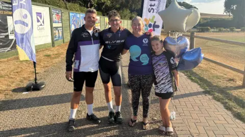 Sarah Brinicombe/BBC Zac after the challenge in Reigate, Surrey, with his family. Zac is at the finish line, and is wearing a purple cycling top. Dad James is pictured to Zac's right, while mum Rachel and sister Bethany are to his left.