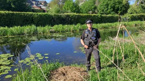 A police officer in dark uniform stood in grass beside a canal with a hedge on the other side
