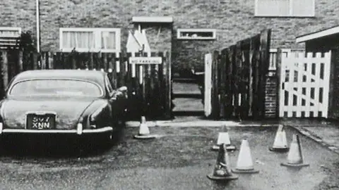 Hertfordshire Police An archive black-and-white image shows a dark saloon car parked outside a row of homes. Several traffic cones are placed on the roadway leading to a narrow fenced pathway between properties. Brick housing and fencing are visible in the background.