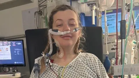 A young pregnant woman in a hospital gown, attached to monitors and in an induced coma