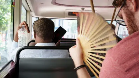 Getty Images The interior of a London bus during a heatwave, showing passengers from behind trying to stay cool. In the foreground, a man in a red shirt rapidly uses a wooden hand fan, creating a motion blur. In the seat ahead, another passenger holds up a cold bottle of water while looking at their phone.