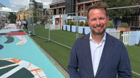 Mark Denten/BBC Stephen Patterson, a middle-aged man wearing a navy pinstripe suit, standing next to the artwork which has more of the elements completed. There is more light blue and a dark green design to the mural.