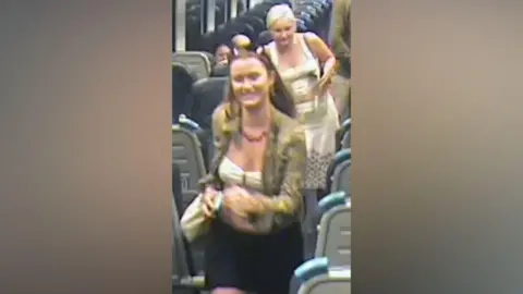 Surrey Police A CCTV image of two women walking inside a train. A woman with brown hair, and wearing a green jacket, white top and black skirt and carrying a bag. A blonde woman, wearing a white dress, is behind her.