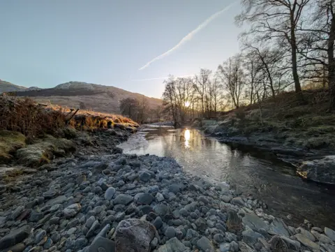 Derek Darkins A partly frozen river on a cold winter's day, with a clear sky above it and hills in the distance