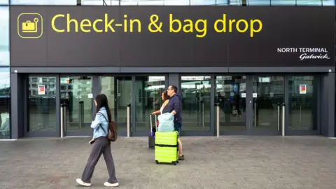 Universal Images Group via Getty Images A sign outside Gatwick airport reads 'check-in & bag drop; north terminal Gatwick', while people walk past wheeling baggage along the concrete floor, image taken in 2024.
