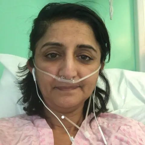 NHS A person lying in a hospital bed wearing a nasal oxygen tube, with medical monitoring equipment visible behind the bed against a pale green wall.