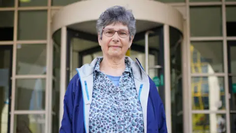 Diana Warner, wearing a blue rain jacket and a patterned blue shirt underneath. She has medium-length black and grey hair and is wearing glasses. She is looking straight at the camera. 