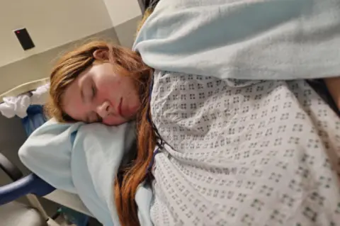 Jade McKenna Sofia-Ann on hospital bed in gown and covered with blanket lying on her side with eyes closed. She has long red hair worn loose.