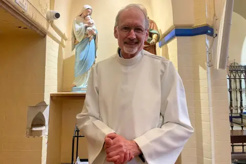 Man smiling, wearing a white vicar's outfit with a dog collar. Behind him is a statue of Mary holding Jesus