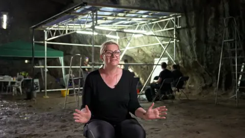 A woman with blonde hair in a black top sits and smiles in front of an archeological dig in a huge cave