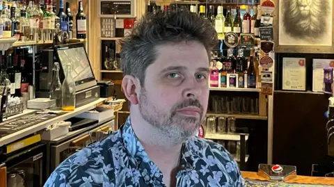 A head and shoulders image of Tom Lagden. He is wearing a black and white floral shirt and sitting in front of a pub bar. 