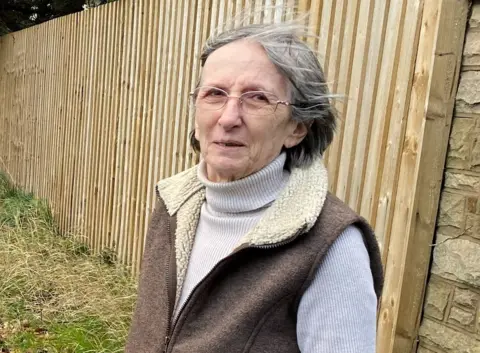 Wendy Pleasent. She has grey hair in a bob and is wearing glasses, a polo neck jumper and sheepskin-type gilet