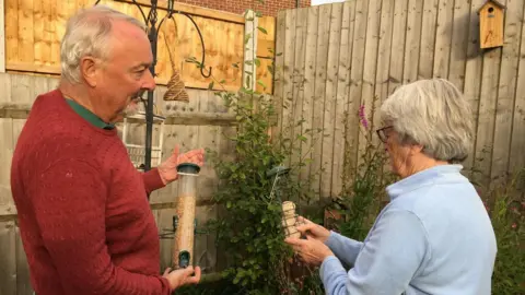 A man with short grey hair is wearing a red jumper. He is holding a bird feeder with seed in it. Behind him is a bird feeding hanger. Next to him is a woman with short grey hair, she is wearing a light blue top.