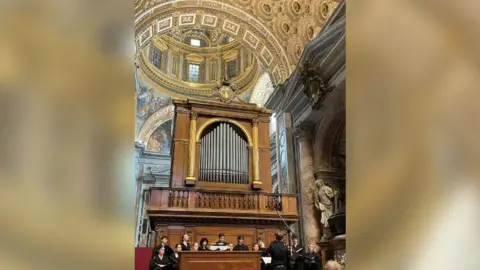 Chapel Choir of University College The choir singing at St Peter's Basilica. They are all dressed in black and there is an organ behind them. The dome is above them.