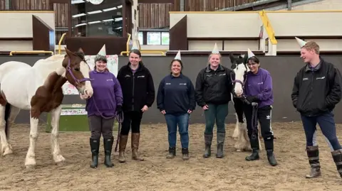 Unicorn Riding Centre A photo from a birthday celebration for one of the horses. The volunteers form a semi-circle in the riding arena, which has a sand floor. They wear boots and party hats. Two of the horses form part of the group. One is a medium-sized horse with brown and white markings. The other is slightly smaller with black and white markings.