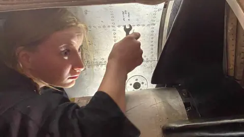 Lightning Association Grace with her hair in a plait, wearing overalls, holding a tool inside the intake of the jet. 