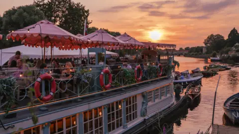 Peggy Jean at Richmond-upon-Thames The Jesus College Barge, which is now Peggy Jean at Richmond-upon-Thames, full of diners on board at sunset. There are some smaller boats floating on the river nearby.