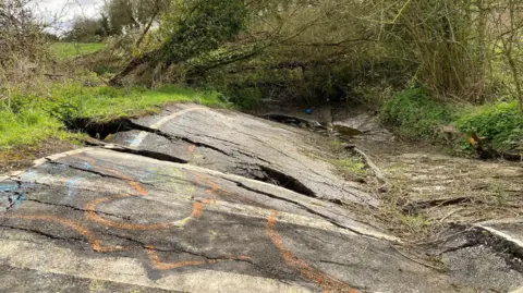 A ripped-up section of road which has been spray painted in orange, blue and yellow paint. The road is no longer a flat, smooth surface and instead goes up and down with dozens of cracks in it of varying sizes.