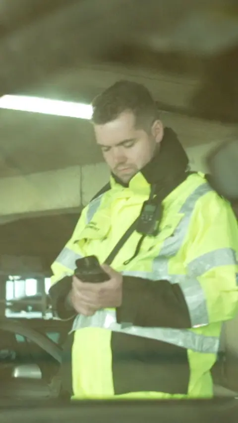 A parking warden in a hi-viz jacket looking at a phone.