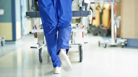 A nurse wearing blue scrubs and white trainers pushes a gurney through a hospital corridor