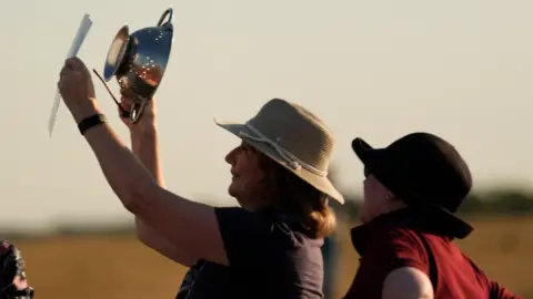 PA Media A woman is holding up a colander and a piece of paper while looking away from the sun