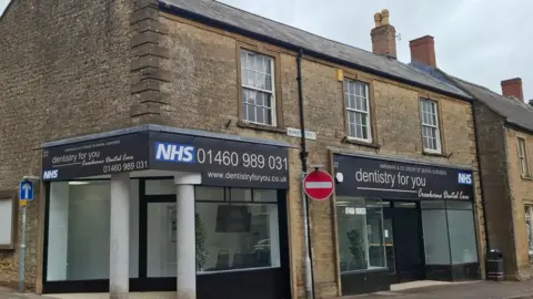NHS The outside of the new dental practice in Crewkerne, which is run by dentistry for you. There are road signs in the image, along with a pavement.