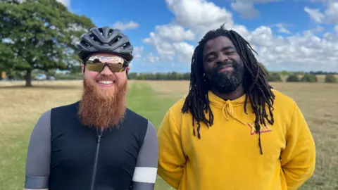 Two men stood in a green field, one wearing cycling gear the other in a yellow hoodie