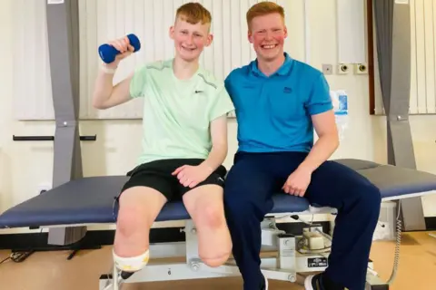 Adam Golebiewski, teenager with both legs removed below the knee, in shorts and a green tee shirt and holding a weight in his right arm, with orthopaedic rehabilitation physiotherapist Owen Cairns, in blue trousers and a blue tee shirt, both sitting on a hospital bed and smiling.