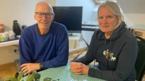 A couple sit at a table with a green tablecloth on it with white spots. She has blonde hair and is wearing a blue hooded top. He is in a lighter blue jumper and has a bald head with glasses.