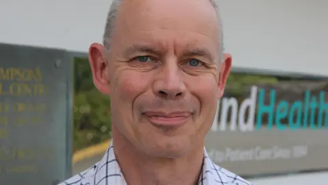 A mostly bald man, with short grey hair on the sides smiles at the camera. He had blue eyes and is wearing a blue checked shirt. Behind him, a shiny sign reads 'IslandHealth', and a plaque says 'St Sampson's Medical Centre.2