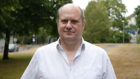 Shaun Whitmore/BBC Prof John Pinnegar a man who is standing outdoors. He is looking directly at the camera and smiling. He is wearing a white shirt and behind him are rows of trees.
