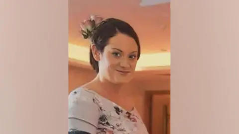 Family handout A head and shoulders picture of Bethanie Clark. She is standing side on to the camera and is wearing a flowery dress. She has her brown hair tied up and has two pink flowers in her hair.