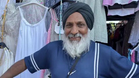 Darren Rozier/BBC A man stands in front of a clothes market stall. He is smiling at the camera. He has a long grey beard and wears a blue polo shirt. He has his hair in a turban.
