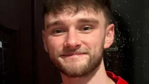 Kieran Thomas Hughes smiling into the camera. he has a short brown beard and brown hair with a fringe. He is wearing a red shirt. 