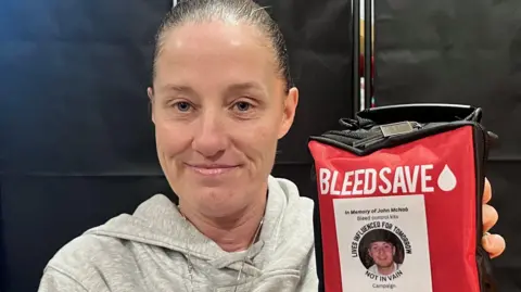 Lisa Petrie has her dark hair tied back and wears a grey hoodie. She holds a red rectangular package which says BLEED SAVE on it. In smaller text it says In Memory of John Mcnab, Bleed control kits, Lives influenced for tomorrow, not in vain campaign