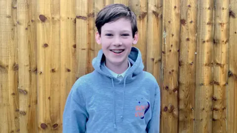 Alex, a young boy with light brown hair, is wearing a grey hoodie and is stood against a wooden fence whilst smiling at the camera.