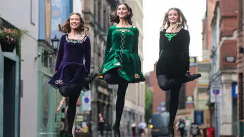 Belfast City Council Three women in traditional Irish dancing dresses - jewel toned velvet with celtic embellishments. They are pictured dancing, mid leap in the air on a city street. 