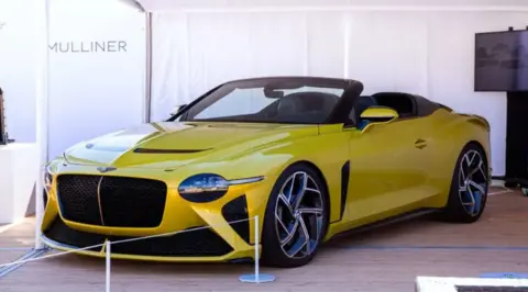 A yellow Mulliner Bacalar on show at a car event. It has its roof down.