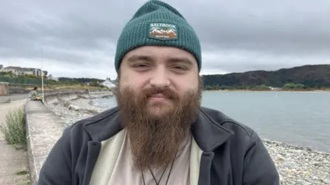 Ryan Shields is wearing a green beanie and a grey jacket. He has a long, light brown beard. 